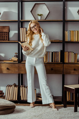 attractive woman in glasses standing near rack and reading book in living room