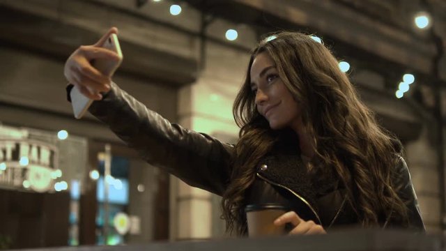 Front view of a woman with a paper coffee cup making selfies in a night city. Concept of social media. Locked down real time medium shot