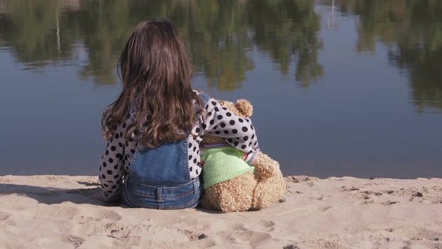 Lonely child with a bear. A little girl on the river embraces a bear cub.