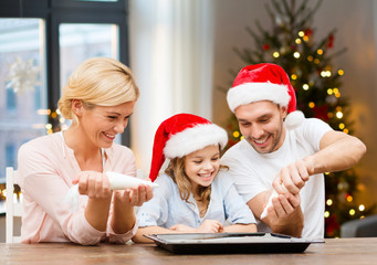 food, family and holidays concept - happy mother, father and daughter family in santa hats decorating bakery by pastry bags with frosting over christmas tree lights background