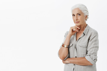 Charming and creative smart senior woman creating new ideas for future business not want retire holding hand on chin in thoughtful pose smiling with delight having idea, thinking over gray background