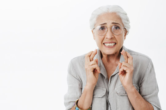 Anxious and concerned hopeful silly senior woman in glasses with white hair crossing fingers for good luck frowning looking intense waiting for impotant news nervously against gray wall