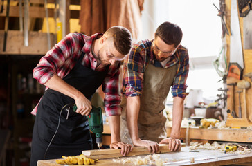 profession, carpentry, woodwork and people concept - two carpenters with electric drill drilling wooden board at workshop
