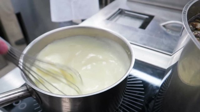 Chef stirring white bechamel sauce in a pan with metal balloon whisk. Slow motion.