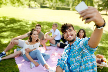 friendship, leisure, technology and people concept - group of friends chilling on picnic blanket abd taking selfie by smartphone at summer park