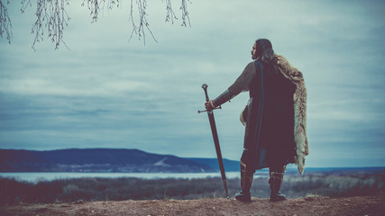 Long haired knight with the two-handed sword. Medieval rural background. - obrazy, fototapety, plakaty