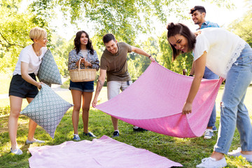friendship and leisure concept - group of happy friends with blanket, pillows and basket arranging...