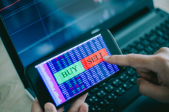 Hand of trader holding mobile phone touch screen showing buy and sell in Stock market order and blurred bakcground of laptop show financial chart, business trading concept