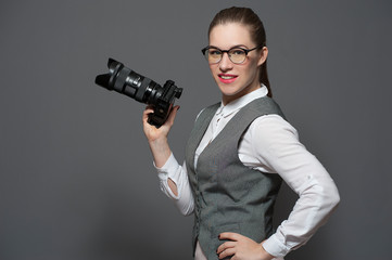 Portrait of smiling confident young woman in eyeglasses dressed in casual clothes her hand on hips holding dslr photo camera standing on grey background