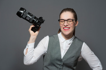 Fototapeta premium Portrait of laughing smiling confident young female photographer in eyeglasses dressed in casual clothes holding a photo camera standing on grey background