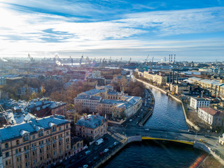 Aerial; drone view of river channel in historical city part with bridges and old house facades near embankment; port with loading cranes and shipyards, wharf on the background; sunny autumn day