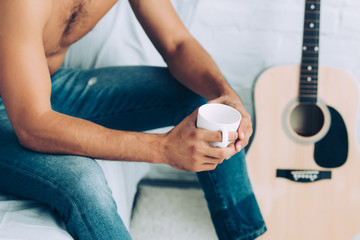 partial view of shirtless man in jeans drinking coffee cup during morning time in bedroom at home