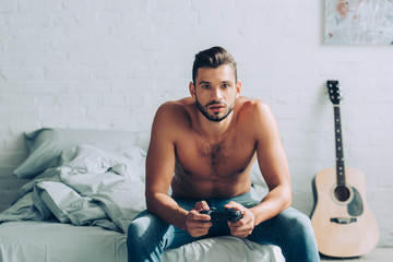 focused shirtless man playing video game with joystick in bedroom at home