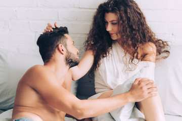 smiling curly woman touching hair of boyfriend in bedroom at home