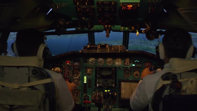 Medium shot from inside of cockpit: male pilots in headsets controlling airplane