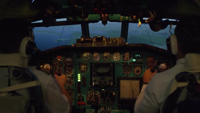 Medium shot from inside of cockpit: male pilots in headsets pressing switches on instrument Panel and controlling airplane with yokes