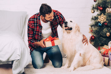selective focus of smiling young man holding christmas gift box near adorable golden retriever at home