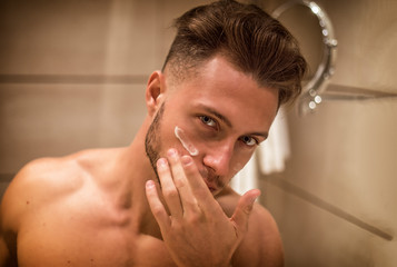 Handsome young man applying moisturizing cream on his face's skin, at home in his bathroom