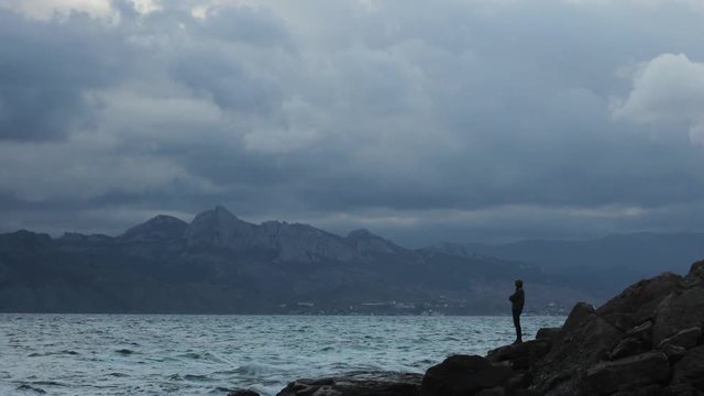 Time Lapse waves hit the shore and man stands at stones Overcast skies move to viewer in front of rocks and capes at day