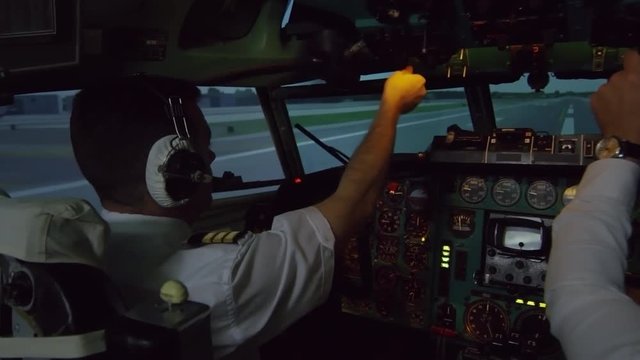 Medium shot from inside of cockpit: male aircraft captain in headset operating switches on overhead instrument panel while controlling airplane moving along runway