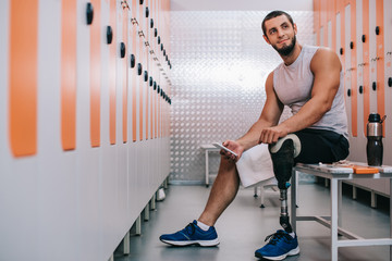 Fototapeta na wymiar smiling young sportsman with artificial leg sitting on bench at gym changing room and using smartphone