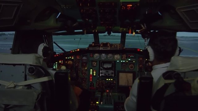Medium shot from inside of cockpit: male aircraft captain and his co-pilot pressing switches on overhead instrument panel and controlling airplane moving along runway
