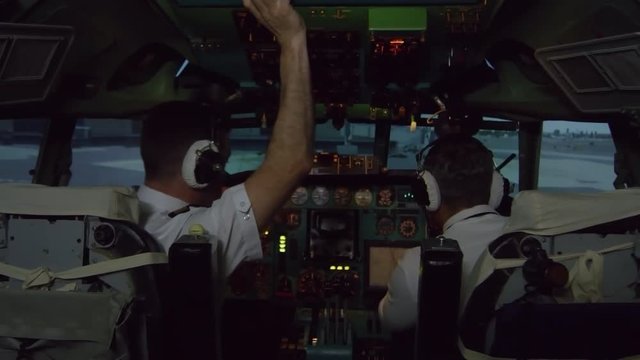 Medium shot of male aircraft captain in headset talking to co-pilot and performing cockpit preparation procedure before flight