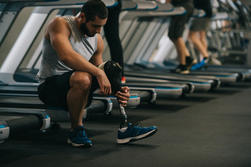handsome young sportsman with artificial leg sitting on treadmill at gym