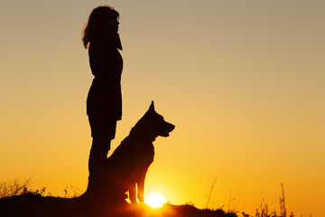 silhouette profile of young woman ang German Shepherddog looking in the distance at sunset, pet sitting near girl's leg on the field, concept of harmony human and nature