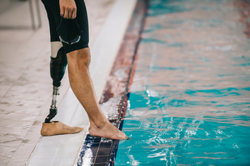 cropped shot of fit swimmer with artificial leg standing on poolside at indoor swimming pool and checking water temperature