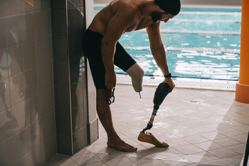 handsome young swimmer standing at poolside of indoor swimming pool and taking off artificial leg