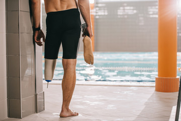cropped shot of athletic swimmer standing at poolside of indoor swimming pool and holding his artificial leg