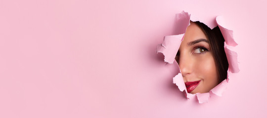 Beauty salon advertising banner with copy space. Beautiful girl looks through hole in pink paper background. Make up artist, fashion, beauty concept. Cosmetics sale. - obrazy, fototapety, plakaty