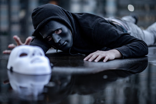 Mystery hoodie man in broken black mask lying in the rain trying to grab white mask on wet floor. Major depressive disorder or bipolar disorder. Depression concept