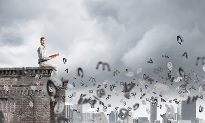 Man on roof edge reading book and symbols flying around
