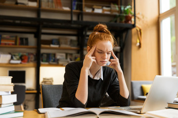 Image of tense concentrated woman studying, while sitting at desk in college library with bookshelf background