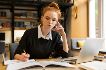 Fototapeta premium Image of intelligent girl studying, while sitting at desk in college library with bookshelf background