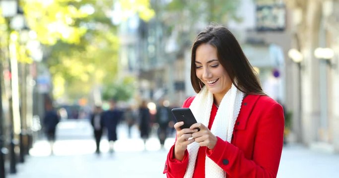 Happy woman wearing a red jacket texting on smart phone in winter in the street