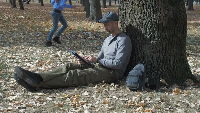 Happy family in the park. Father with daughters in the autumn park. A man with a tablet sits under a tree.