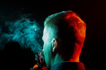 A view from the back of a guy’s head around in a shirt smoking a vape and exhaling multi-colored smoke of green to the left side from himself on a black isolated background. Harm to health of human.