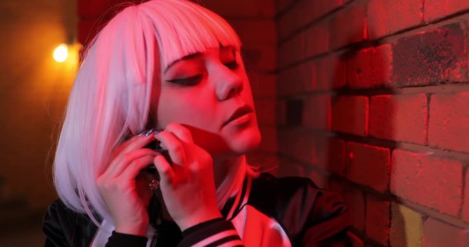 Sexy unusual girl in white wig fastens big earring, red light brick wall
