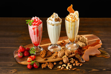 Three sweet milkshakes with nuts, caramel, strawberry and whipped cream at a wooden board on table...