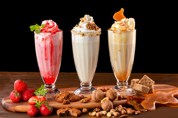 Three sweet milkshakes with nuts, caramel, strawberry and whipped cream at a wooden board on table...
