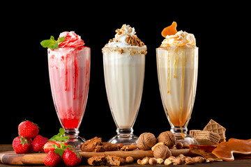 Three sweet milkshakes with nuts, caramel, strawberry and whipped cream at a wooden board on table background. - obrazy, fototapety, plakaty