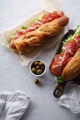 Top view of two fresh baguette sandwiches bahn-mi styled. Ham, sliced cheese, tomatoes and fresh lettuce on dark wooden cutting board on concrete background.