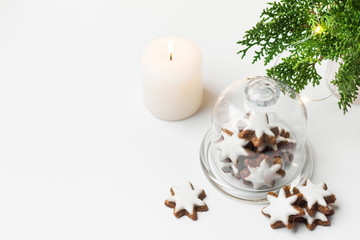 Traditional German star shaped cinnamon cookies with icing in cloche dome bell glass jar on white table. Lit candle juniper with golden garland lights. Magic festive atmosphere