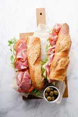 Fresh baguette sandwich bahn-mi styled. Ham, sliced cheese, tomatoes and fresh lettuce on wooden cutting board on white marble background.
