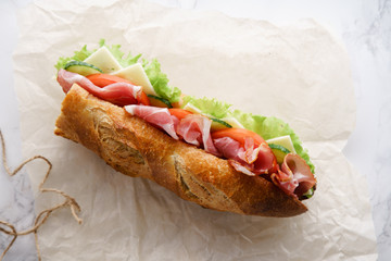 Fresh baguette sandwich bahn-mi styled. Ham, sliced cheese, tomatoes and fresh lettuce in wrapping paper on white marble background. Top view, copy space