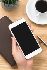 Male hand holding smartphone with notebooks, pen, and coffee cup beside on wood table in morning time