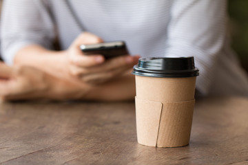 Paper coffee cup hot drink on wooden desk with woman working on smart-phone blurred in background